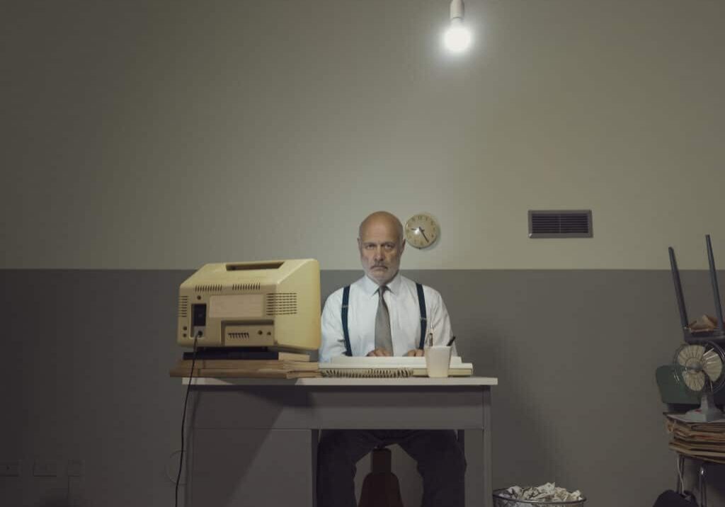 Sad vintage style businessman working in a small office with an old computer and cheap furniture, he feels frustrated and disappointed