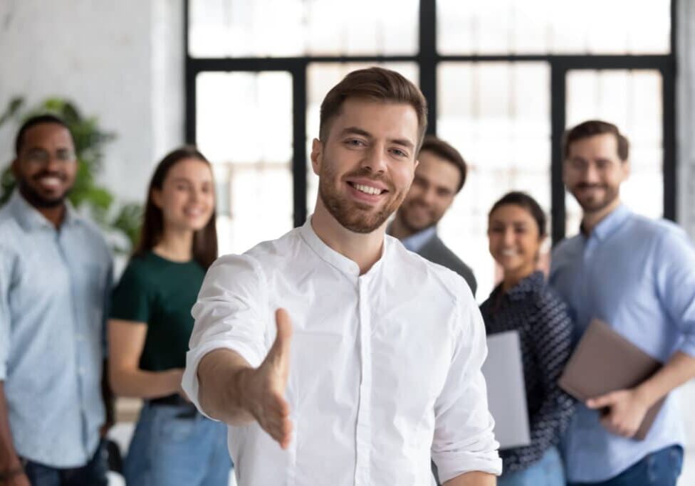 Smiling young Caucasian businessman stretch hand greeting meeting with new office employee or worker, happy male team leader or employer welcome newcomer or intern at workplace, recruitment concept