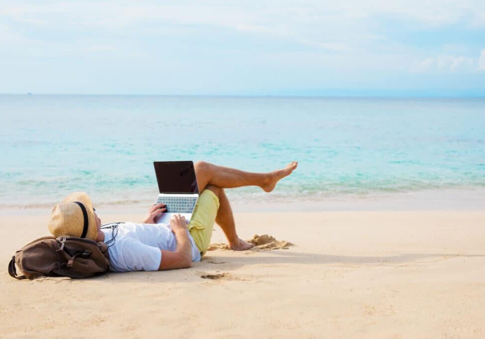 Man working on laptop computer while relaxing on the beach. Idyllic photography covering working anywhere concept.