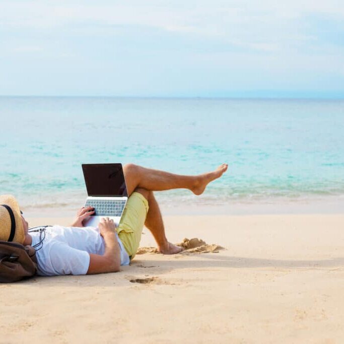 Man working on laptop computer while relaxing on the beach. Idyllic photography covering working anywhere concept.
