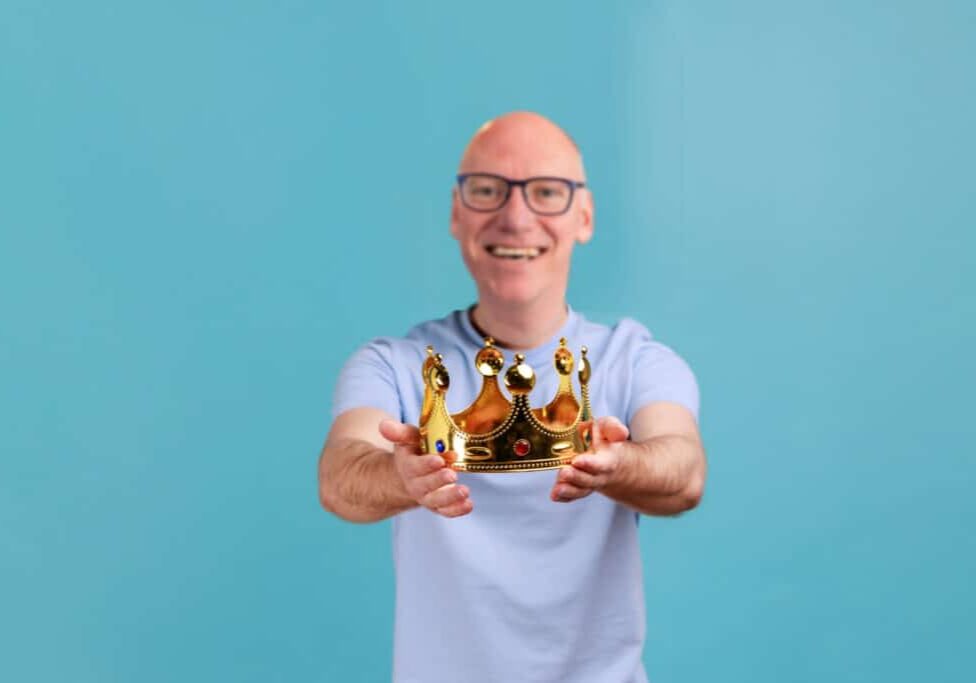 Man holding out golden crown, looking at camera with toothy smile, expressing positive emotions. Portrait of smiling happy bearded man holding out golden crown, looking at camera with toothy smile, expressing positive emotions. Indoor studio shot isolated on blue background.