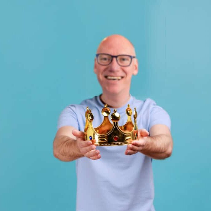 Portrait of smiling happy bearded man holding out golden crown, looking at camera with toothy smile, expressing positive emotions. Indoor studio shot isolated on blue background.